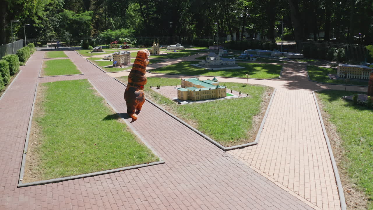 Person in air costume of dinosaur walking along the miniature buildings. Tirex creature having a walk between the outdoor museum on sunny summer day.