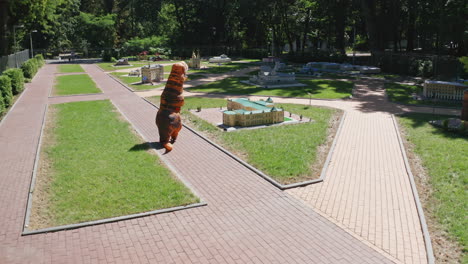 person in air costume of dinosaur walking along the miniature buildings. tirex creature having a walk between the outdoor museum on sunny summer day.