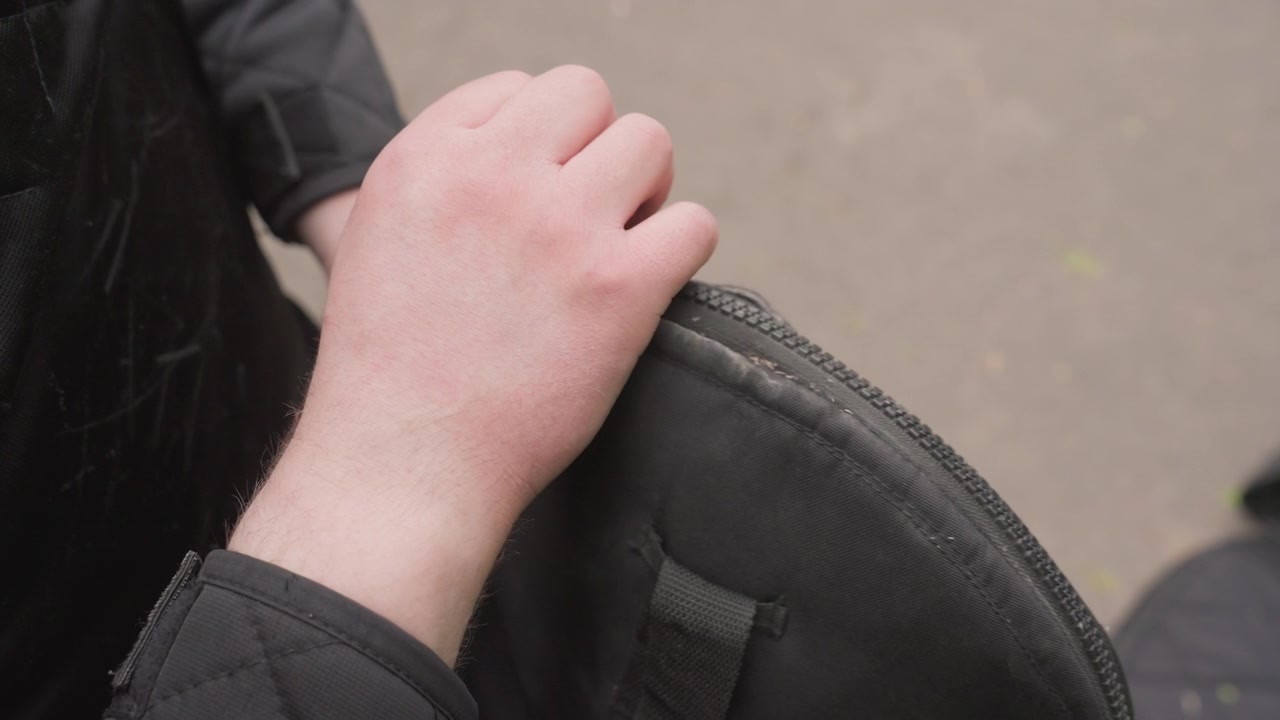 Primer plano de persona manipulando y abriendo un bolso acolchado negro con costuras y cremallera visibles. Las manos de la persona interactúan con la tela revelando detalles de la textura mientras lo desabrochan. Fondo