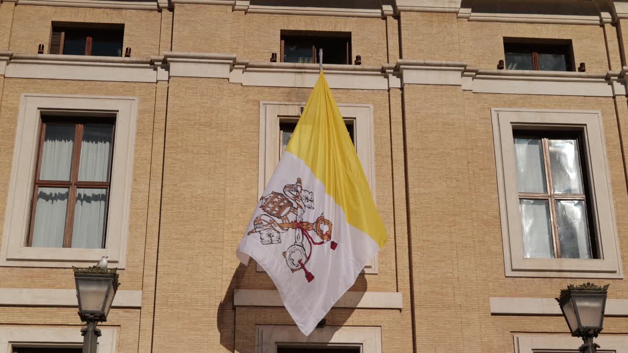 The Vatican City flag waves gently in the breeze in front of a historic Roman building, captured in real-time under bright daylight with soft camera motion.