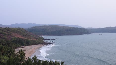 A-serene-beach-in-Goa-with-coconut-trees,-waves,-and-distant-hills-during-the-day