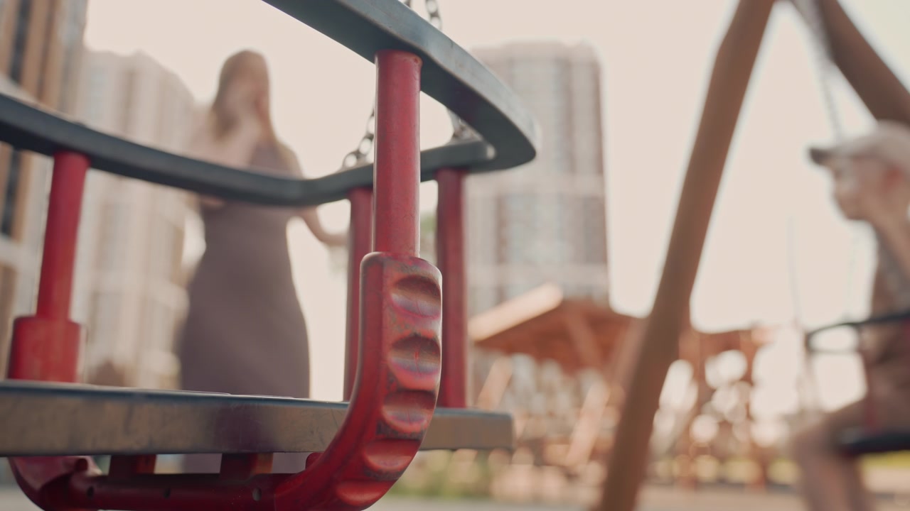 Una donna e un bambino interagiscono in un parco giochi urbano, con il primo piano che mostra componenti di altalene in metallo rosso. Lo sfondo presenta figure sfocate sulle altalene contro edifici alti sotto una luce diurna soffusa.