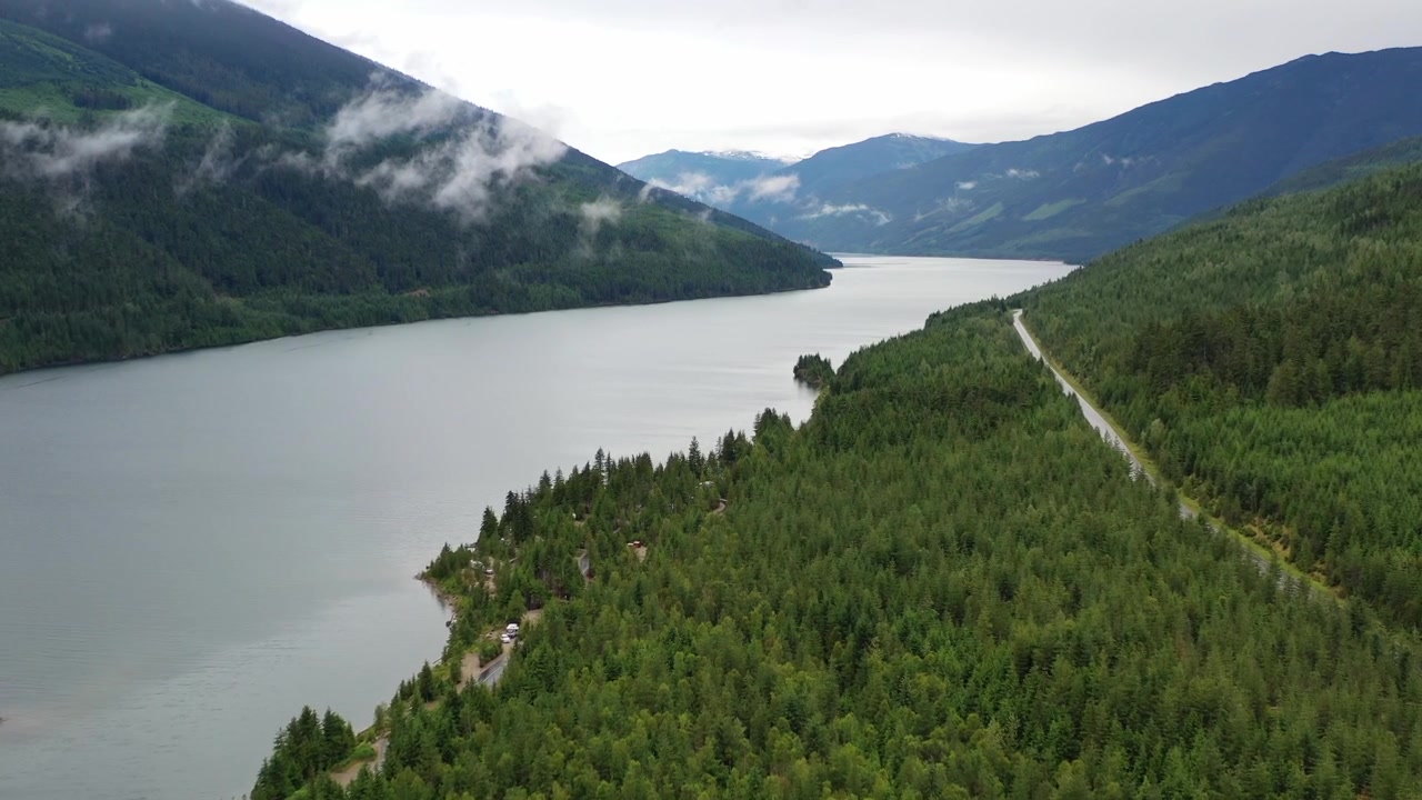 Ripresa cinematografica con drone dall'alto di una strada stretta che si snoda attraverso una fitta foresta di conifere accanto a un lago calmo e riflettente. Cime montuose suggestive e nuvole basse definiscono il paesaggio della Columbia Britannica.
