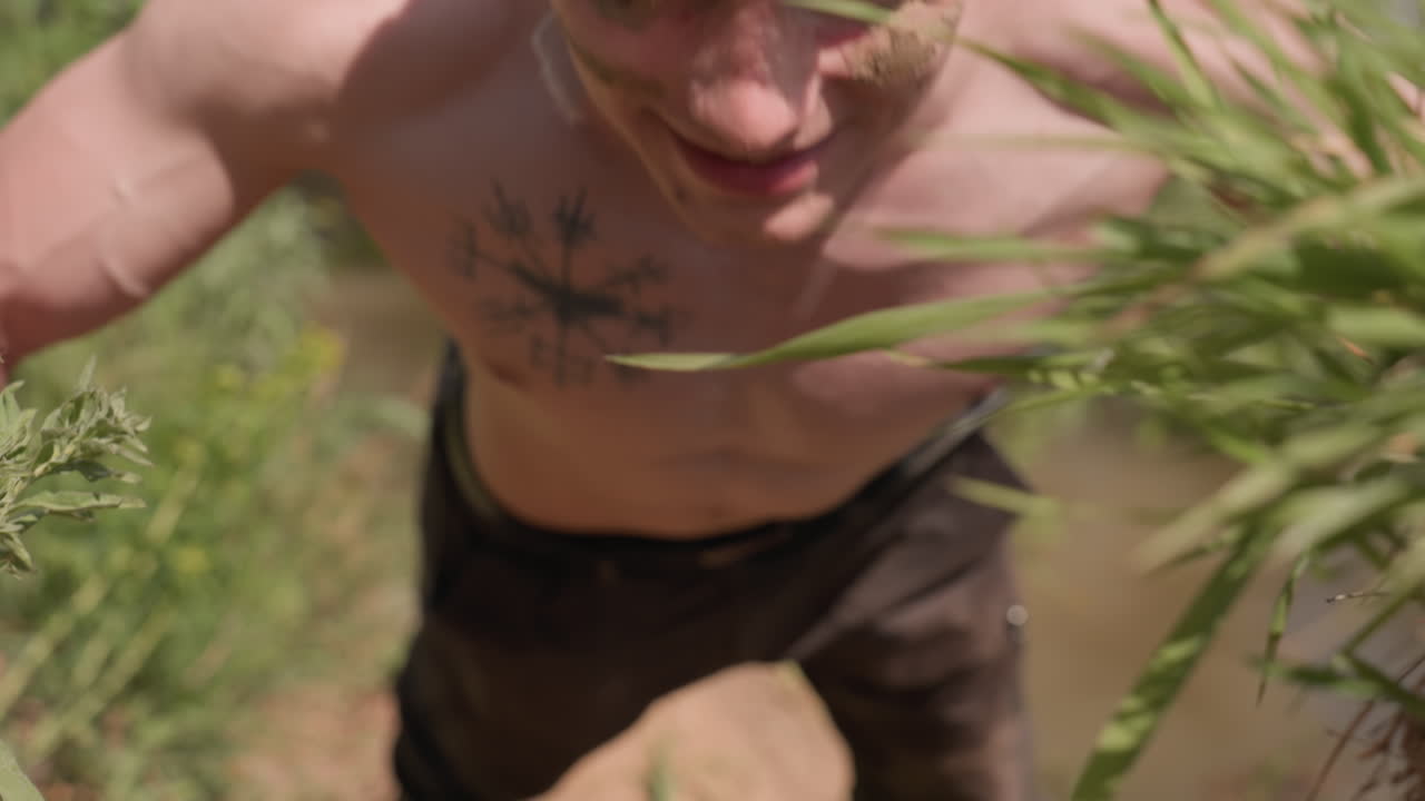 close view of fighter pushing up with veins bulging on chest, sand camo on face, wrap, shirtless young man training on grassy bank dirt slope by water under sun, muscles flexed with effort