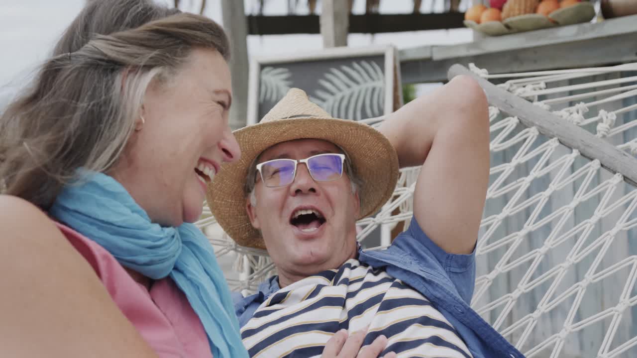 una pareja caucásica senior riendo relajándose en una hamaca fuera de la casa de la playa, en cámara lenta