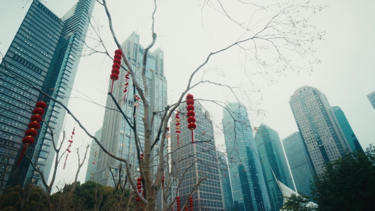 Plano ascendente de lanternas vermelhas do Ano Novo Chinês penduradas em galhos nus de árvores de inverno em Lujiazui Central Green, Xangai, com os arranha-céus do distrito financeiro de Lujiazui elevando-se através de uma neblina pálida de inverno