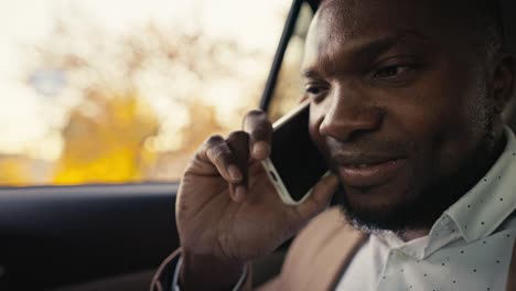 close up of a happy man businessman with black skin color in a brown jacket communicates on a smartphone while riding in the back seat of a modern car outside the city during his business trip