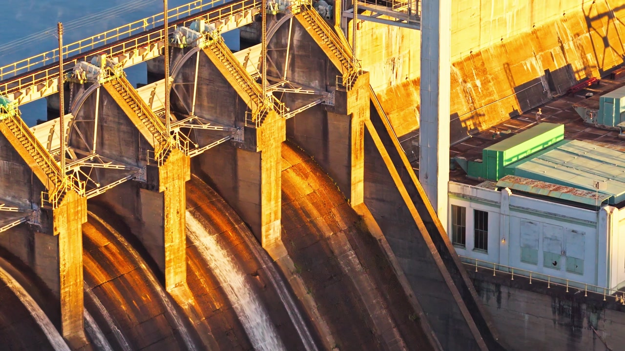 Golden sunlight illuminating spillway gates and concrete structure of hydroelectric dam, aerial backdrop with engineers