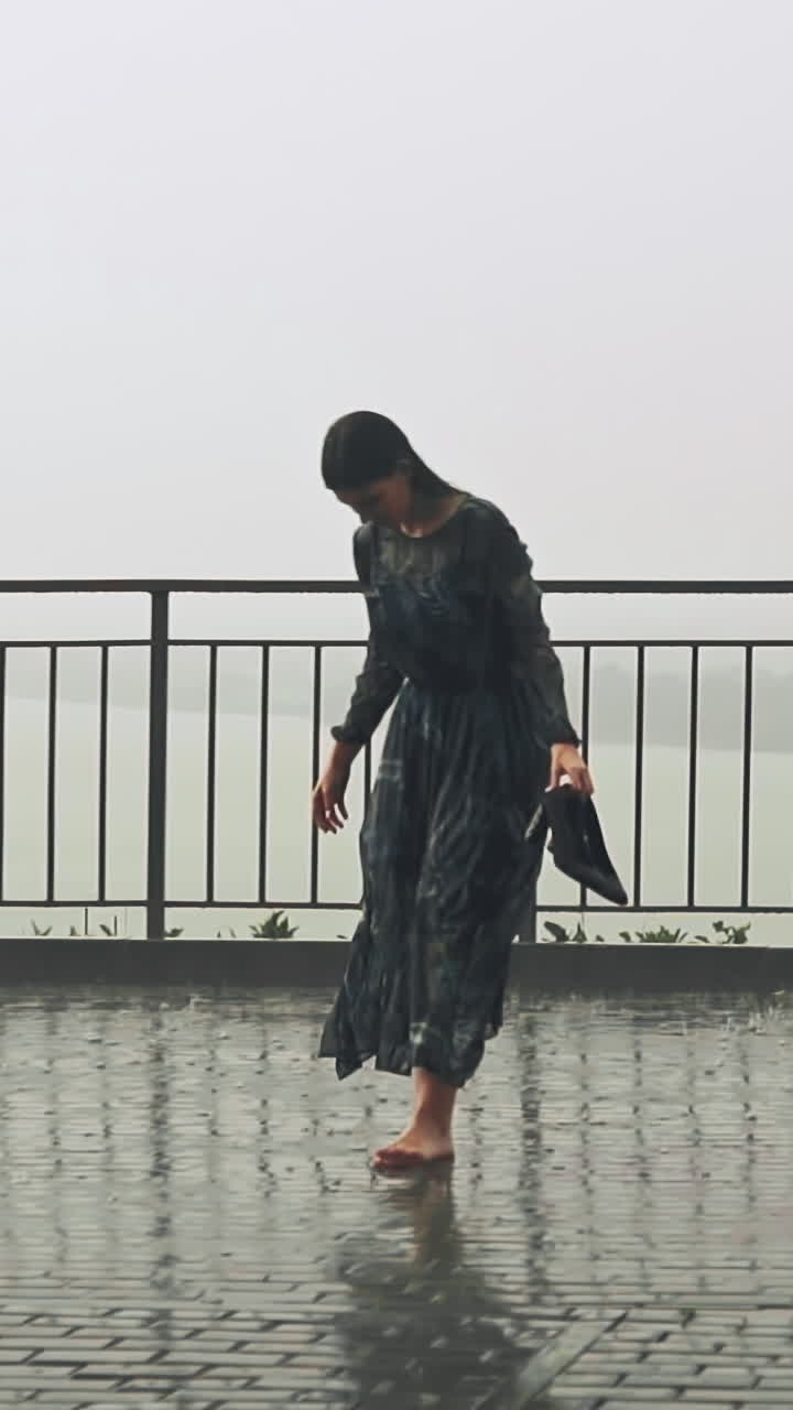 Funny barefoot young woman in wet dress stands holding heeled shoes in puddle under heavy rain on viewing deck against foggy city on spring day slow motion