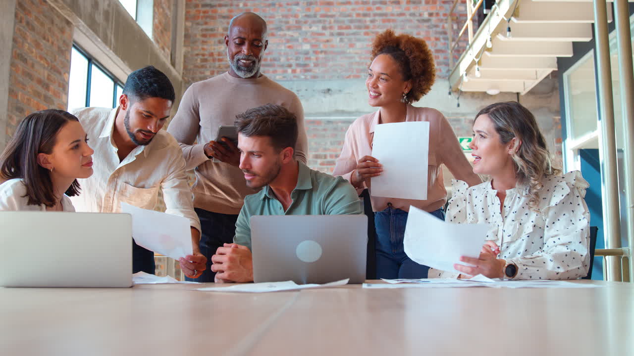 Multi-Cultural Business Team Meeting Around Laptop Discussing Documents In Busy Office