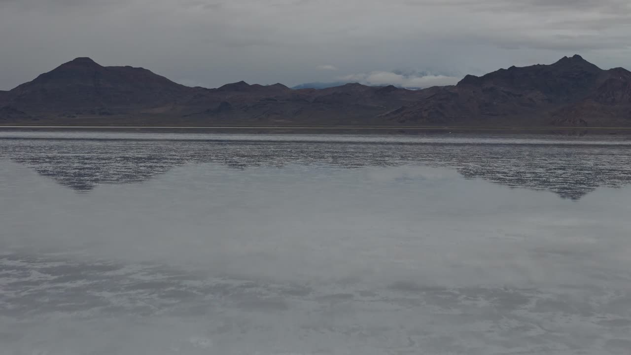 drone lenta aproximación baja sobre bonneville salinas con agua que refleja el cielo y nubes con montañas en el fondo