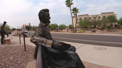 Primer-Plano-Medio-De-La-Estatua-De-Bronce-De-Abraham-Lincoln-Sentado-En-Un-Banco-Del-Parque-Con-Vistas-A-La-Carretera-Principal,-Fountain-Hills,-Arizona