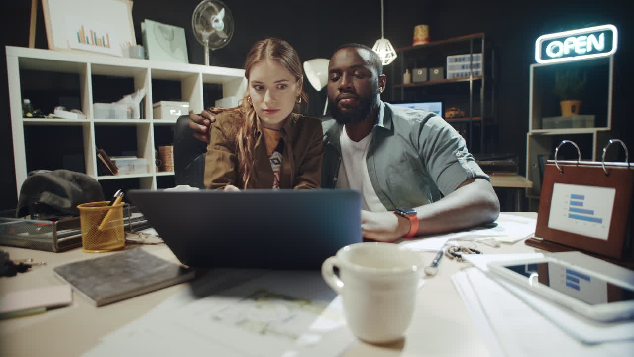 African american man hugging surprised woman near laptop in dark office.