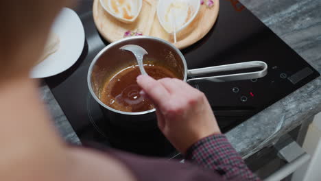 partial view of person stirring thick soup in stainless steel pot on induction stove while adjusting its position, with wooden tray, used bowls, and kitchen utensils arranged on dark marble