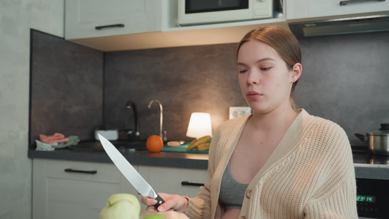 Pregnant caucasian woman preparing food at kitchen counter, chopping green apples with knife, focused expression, visible baby bump, emphasis on prenatal nutrition, simple home cooking, warm lamp