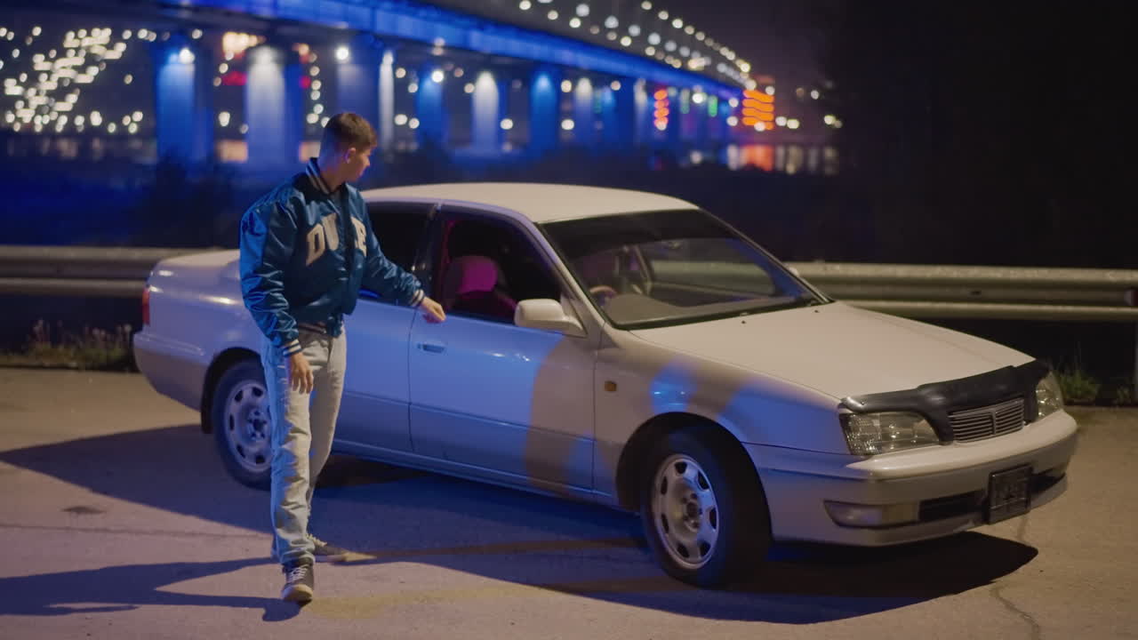 주차된 차에 앉아 있는 백인 남성, 파란 다리 조명 아래, 열린 문에서 몸을 기울인 운전자의 도시적인 야간 초상화, 핑크색 내부 조명, 자신감 있는 자세, 무릎에 올린 손, 영화 같은 색 보정