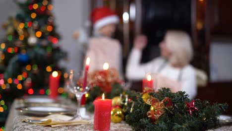 close-up of christmas candle burning on decorated table with blurred boy putting on new year hat and woman showing thumbs up at background. relaxed caucasian family on holiday at home.