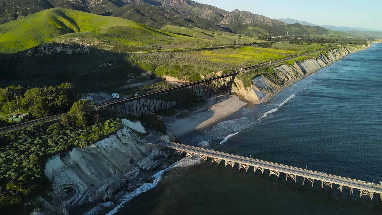 Aerial establishing dolly of ocean waves crashing on sandy beach with empty pier and railroad trestle with green hills, Gaviota Beach, California
