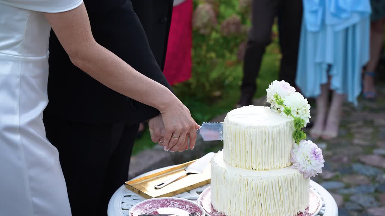 Um casal corta elegantemente um bolo de vários andares lindamente decorado durante uma cerimônia de casamento ao ar livre. A iluminação natural suave realça o design branco e floral do bolo.