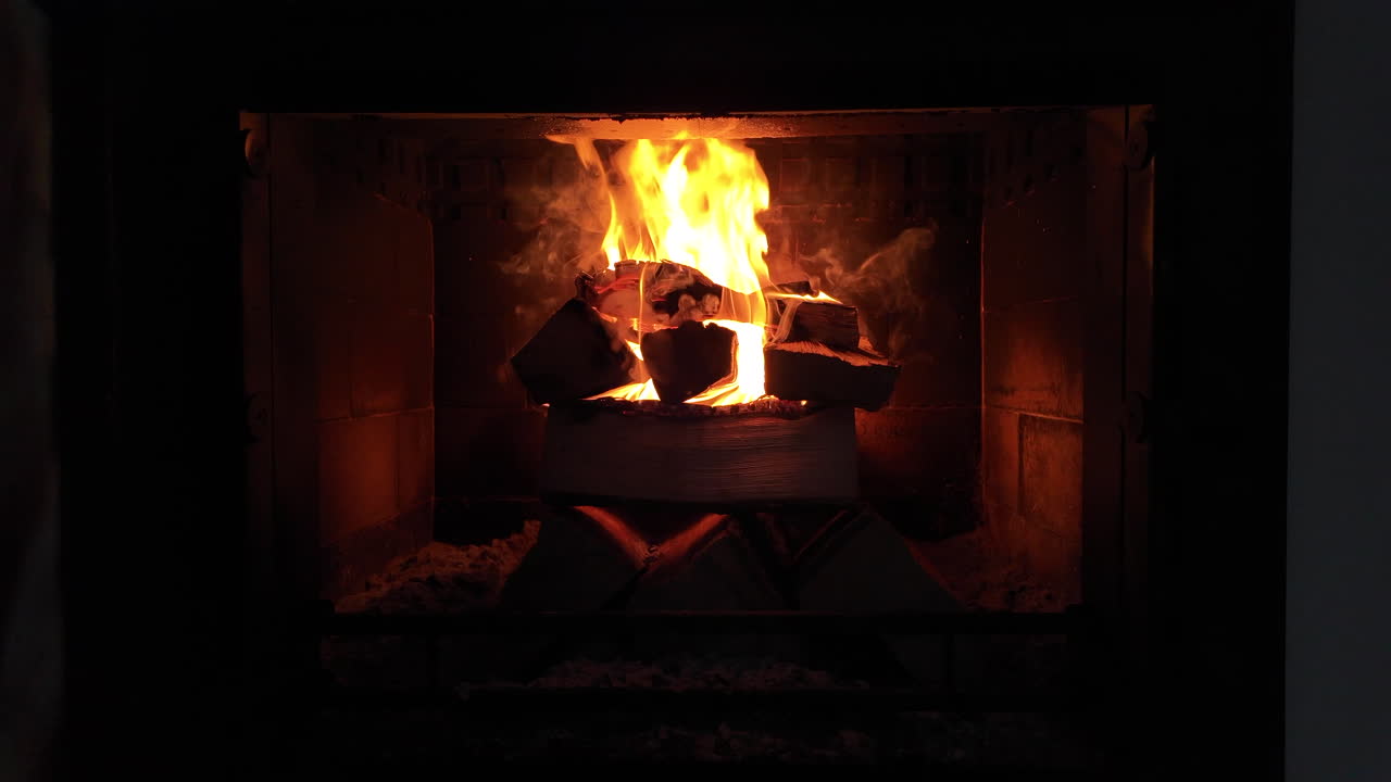 A steady fire burns inside a traditional brick fireplace, with glowing flames rising around stacked firewood in a dark interior. The warm light creates a cozy and intimate atmosphere