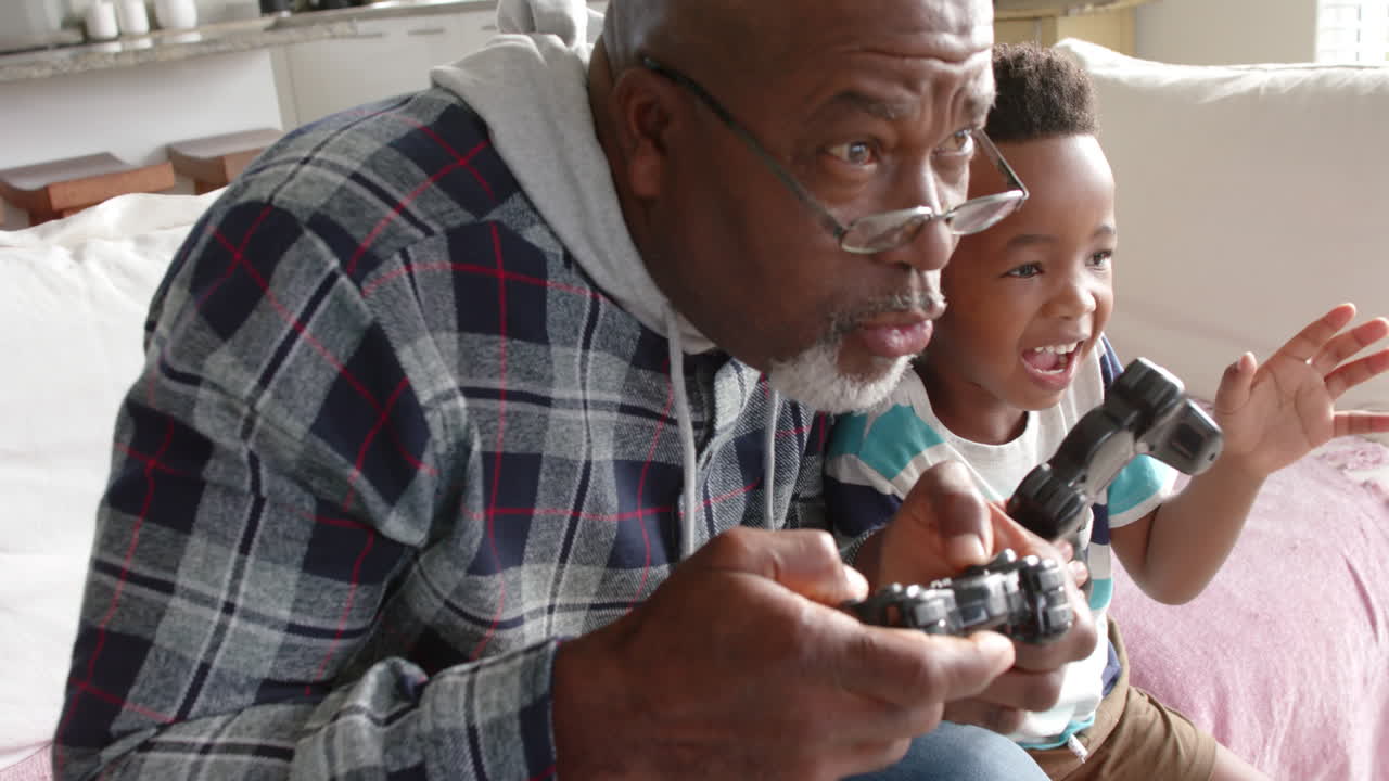 African american grandfather and grandson sitting on sofa and playing video games, slow motion