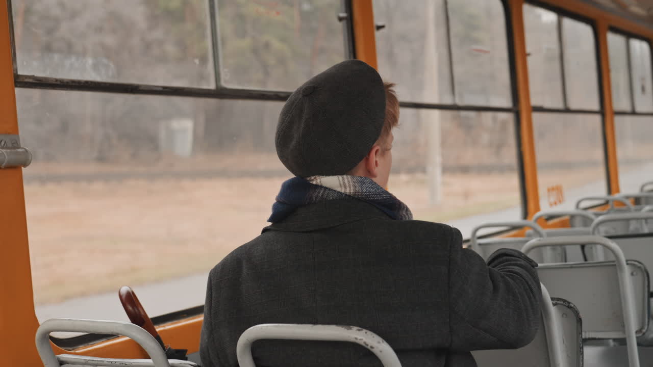 Creatore anziano bianco seduto su un tram, che guarda attraverso il finestrino appannato un campo autunnale, indossando basco e sciarpa, ombrello accanto al sedile; file vuote e interni vintage dai toni tenui creano un'atmosfera contemplativa.