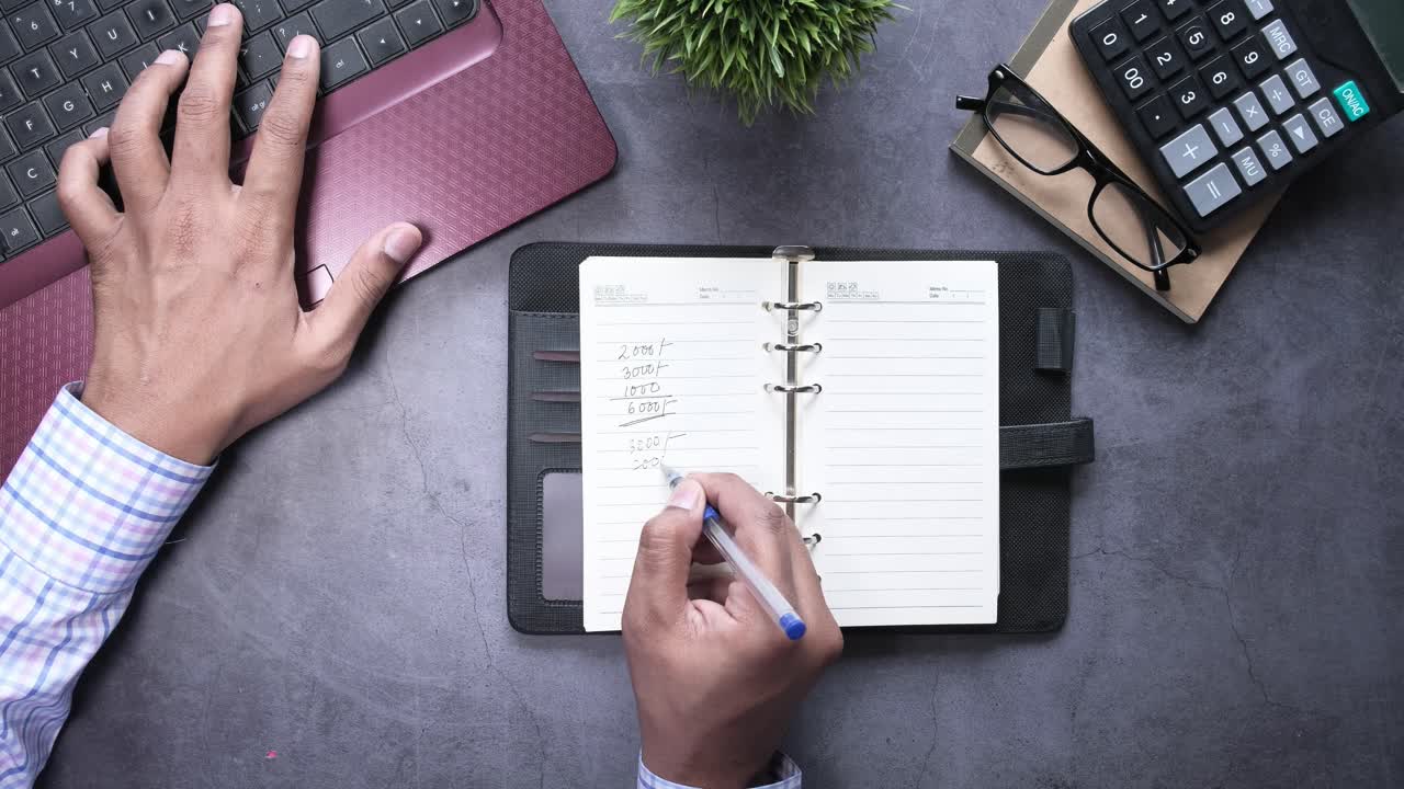 Person writing in a notebook at a desk with a laptop