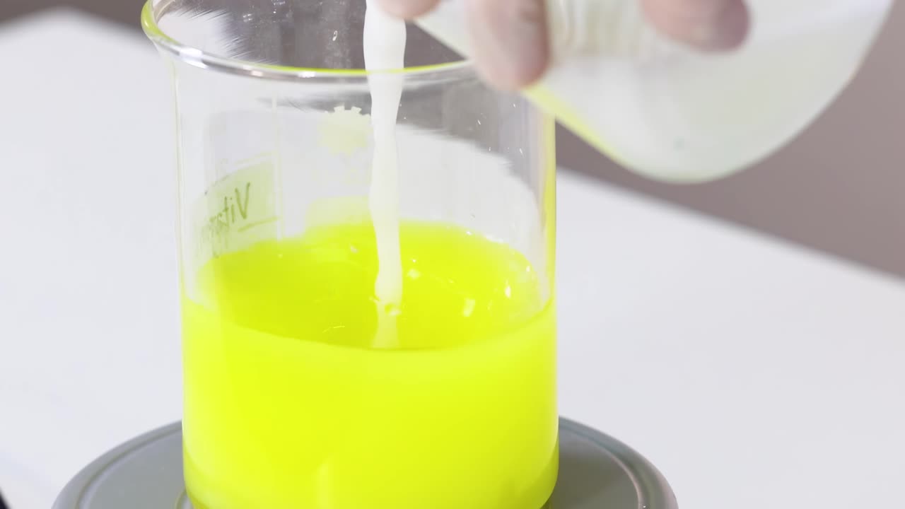 Se vierte un líquido en un vaso de precipitados que contiene una solución amarilla brillante, demostrando un proceso químico.