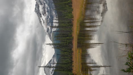 vertical time lapse, cold autumn day in mountains of canada, dramatic dark clouds above snow capped peaks and valley with conifer forest and lake water