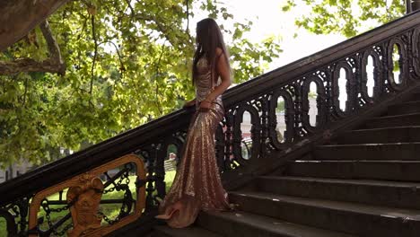 Attractive-young-fit-woman-in-a-long-golden-shiny-dress-walks-down-on-the-stairs-on-high-heels-in-summer.-Party-preparation