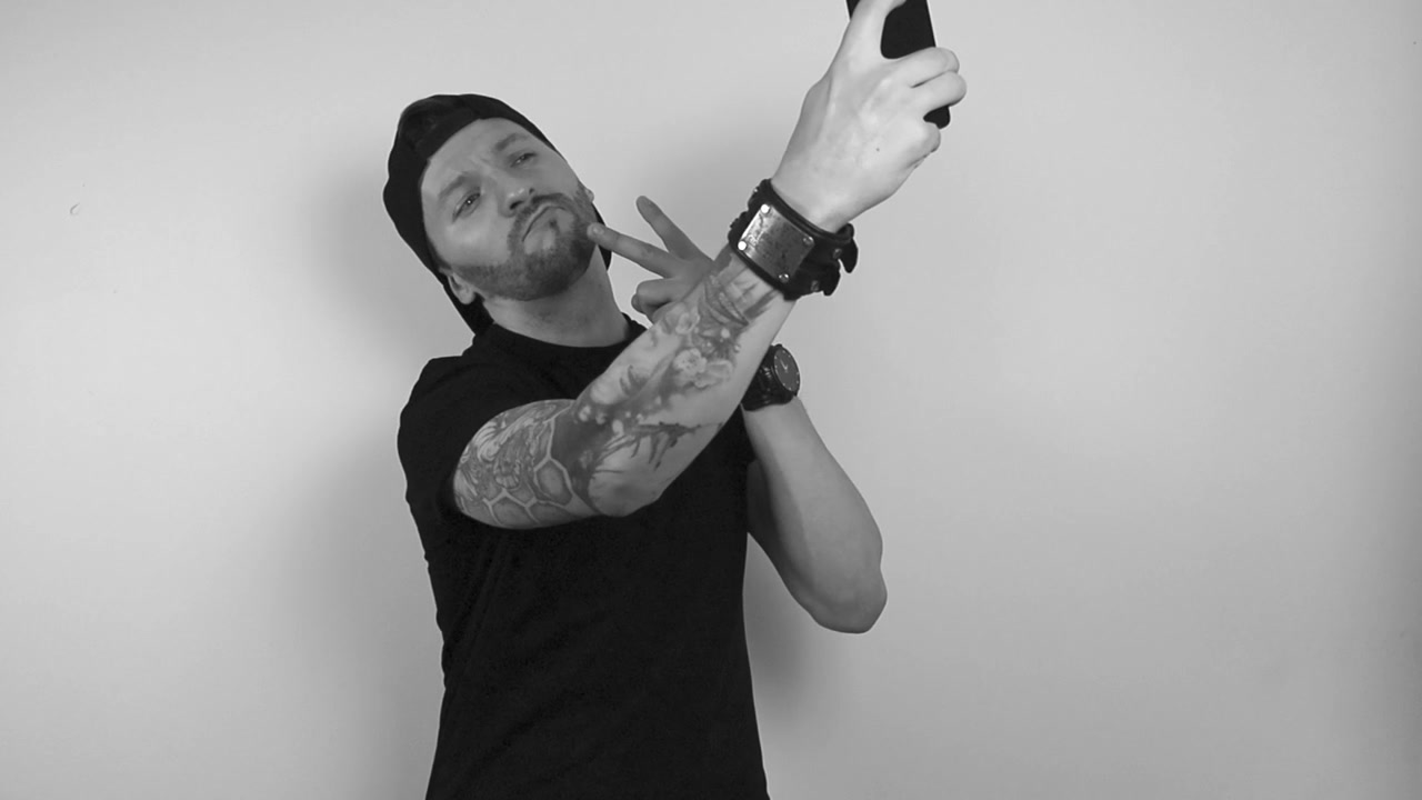 Riprese in bianco e nero di un uomo tatuato in posa davanti a uno smartphone o a una fotocamera del telefono, mentre si scatta un selfie e mostra il segno della pace con l'altra mano. L'uomo appare compiaciuto e felice di pubblicare sui social media.