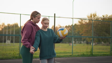 volleyball instructor teaching beginner how to serve on outdoor court with volleyball in hand, instructor demonstrating technique with distant football field and goalposts in background