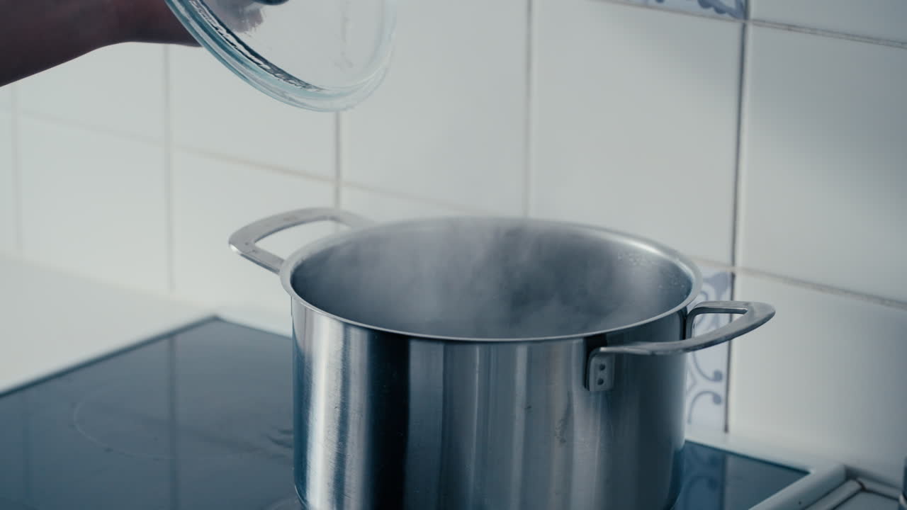 Close up of woman's hand opening the Lid on a big Iron Pot with boiling and steaming Water bubbling on the stove in the kitchen in slow motion