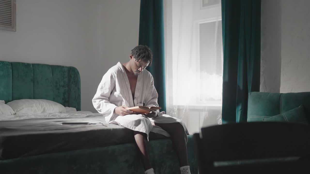 Guy in bathrobe sitting on bed, reading book in bright morning light. Soft sunlight illuminates room, creating a tranquil, cozy atmosphere. Peaceful moment in comfortable indoor space