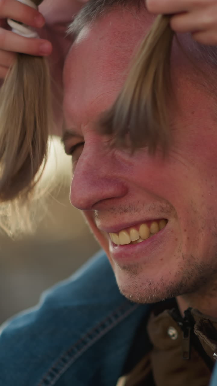 una niña con una gorra y una chaqueta rosadas se sienta alegremente en el cuello de su padre mientras caminan a través de un parque iluminado por el sol. ella juega inclinando la cabeza hacia abajo y usa su cabello para jugar en su cara