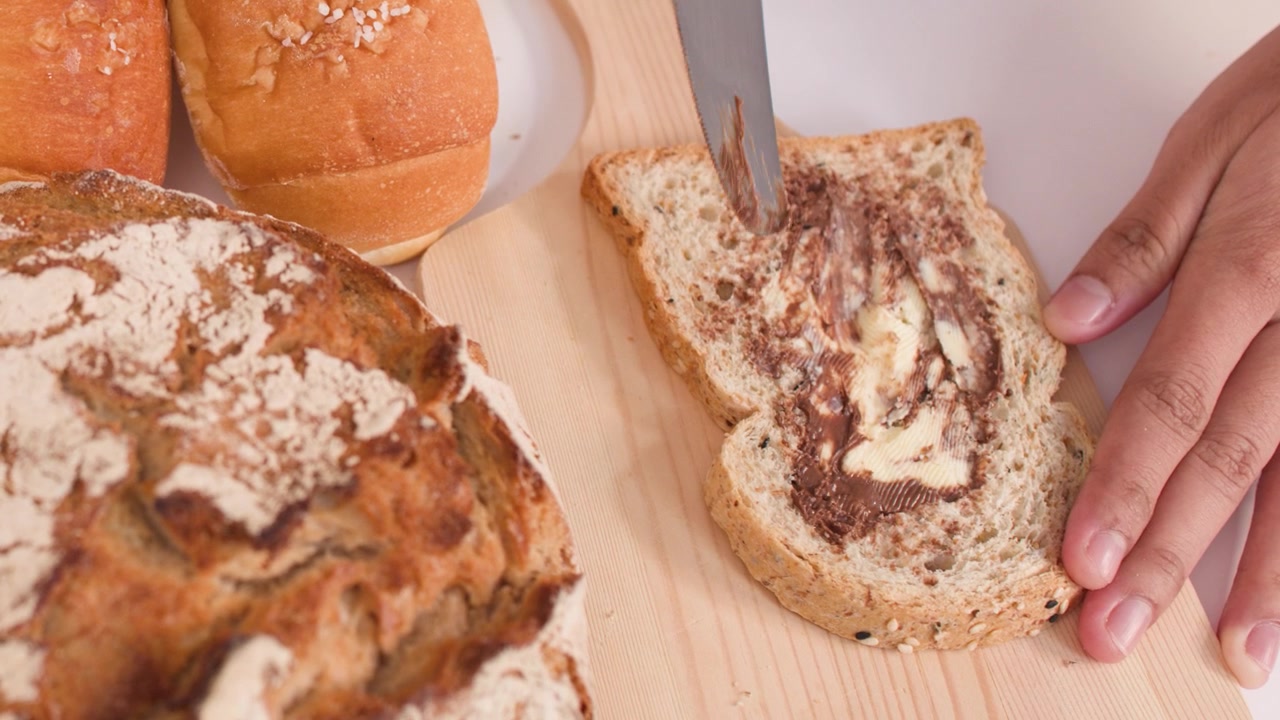 Close-up of hands spreading peanut butter on bread amidst sourdough, baguettes, and croissants.