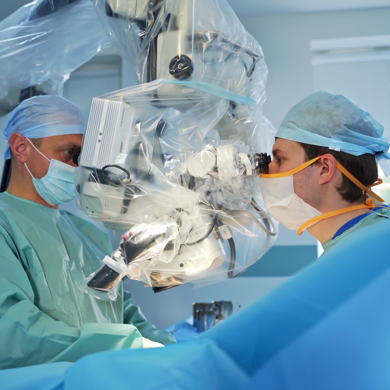 Couple of surgeons stand on both sides of modern equipment looking into binoculars. Hands of doctors with metal instruments move under the apparatus dealing with patient's body