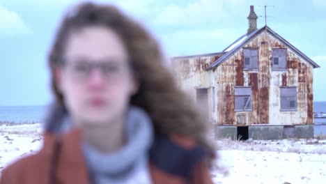 camera switches focus between closeup portrait of young red hair girl and an abandoned, rusty house in the background.