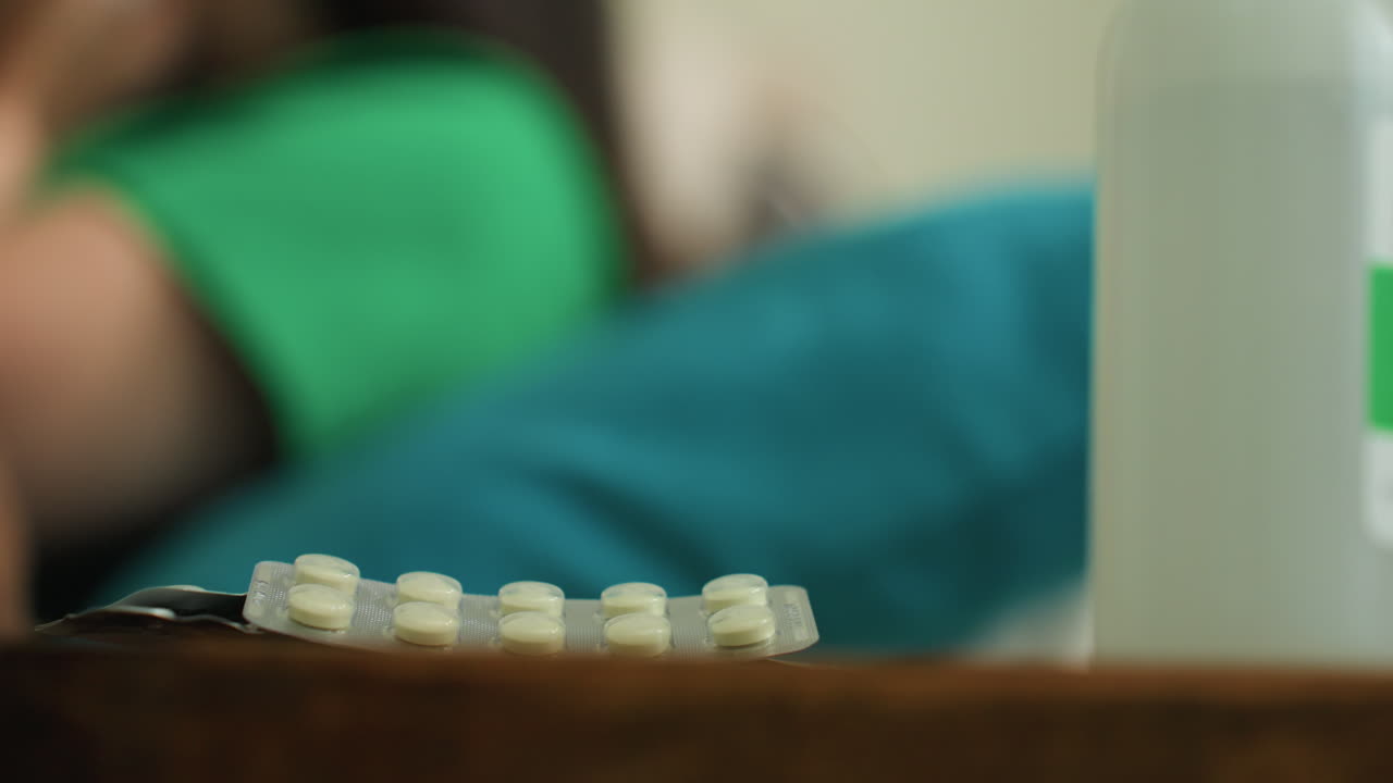 실내에 약이 놓여 있고, 알약에 초점이 맞춰져 있으며, 뒤에는 여성이 흐릿하게 보입니다. 가정집 테이블에 약이 놓인 모습으로, 배경에서 여성이 휴식을 취하고 있고 테이블 위에 약 알약이 놓인 사진입니다.