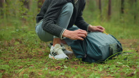 partial view of person squatting in forest while unzipping camping gear bag placed on grass with slight view of another bag nearby showing outdoor activity and preparation in natural woodland setting