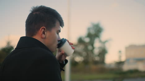 man in black coat sipping tea while deeply focused in an outdoor setting featuring blurred background of greenery and building, under soft lighting