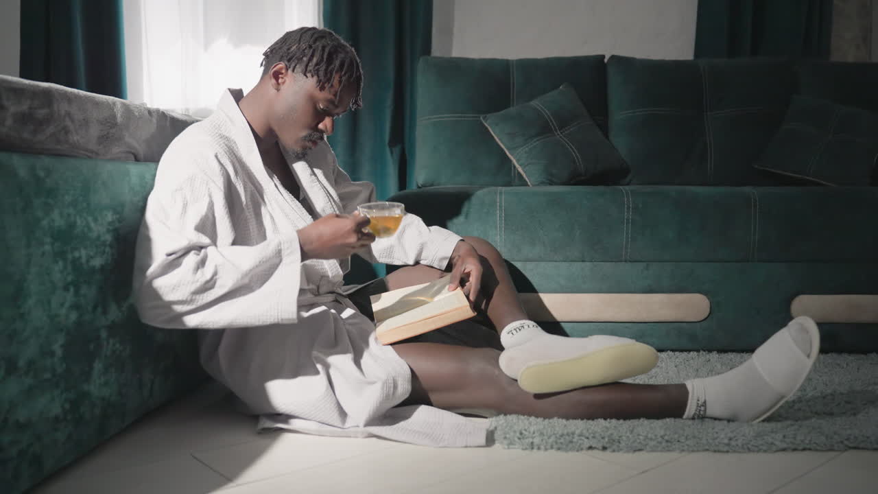 Man in white robe sitting on rug, reading book while holding tea cup. Bright morning light shines on modern bedroom with green furniture, creating relaxing and peaceful atmosphere for self-care