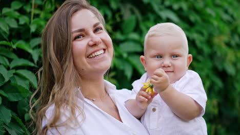 happy smiling caucasian mom with her infant baby outdoors. cute smiling boy is holding a yellow flower. close up.