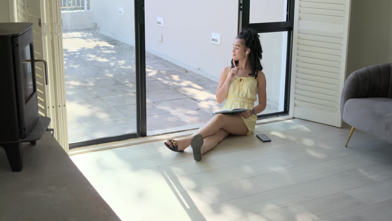 Mujer afroamericana sentada junto a una puerta corrediza de cristal, tomando notas en un cuaderno mientras suena el teléfono