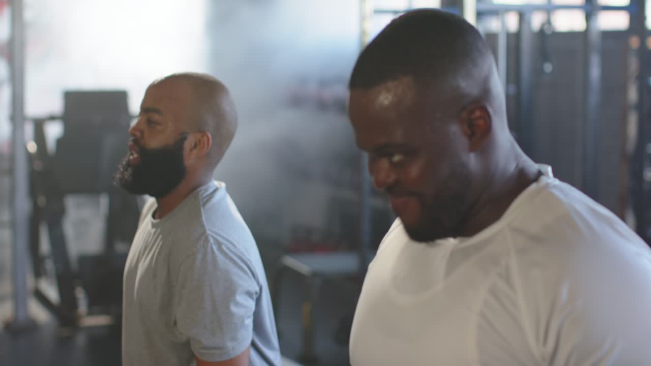 Two African men in T-shirts curling hex dumbbells at gym, right man starting set, building strength