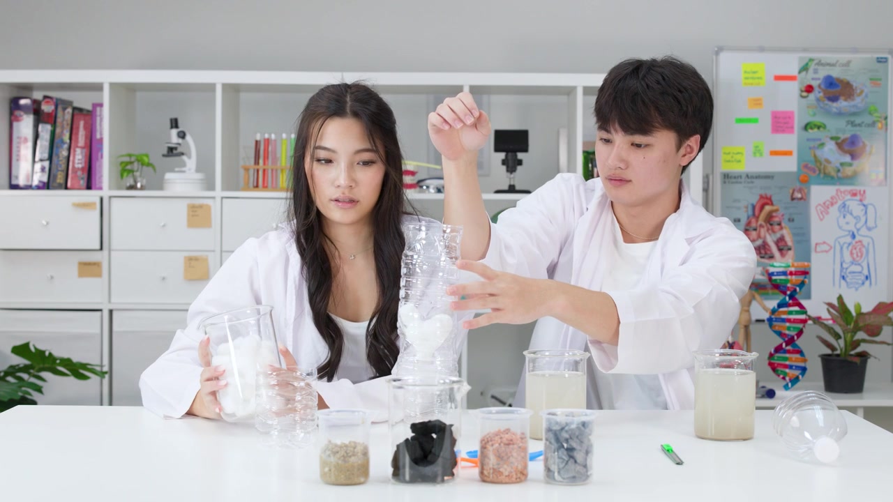 Dos estudiantes con batas de laboratorio ensamblan un filtro de agua casero usando botellas de plástico y algodón