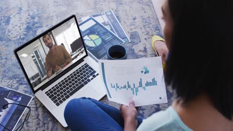 Mixed-race-businesswoman-sitting-on-floor-using-laptop-having-video-call-with-male-colleague