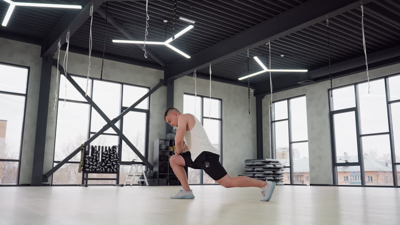 Young man stretching feet and legs with forward lunge in bright gym room, wearing white tank top and black shorts, large windows and equipment racks in background, showcasing warm up, and strength