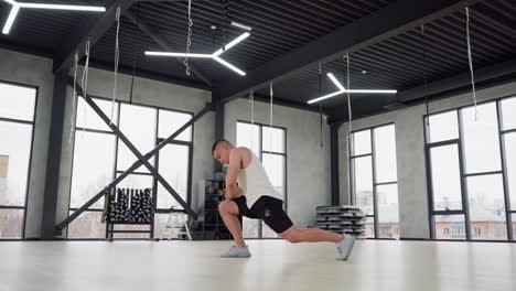 young man stretching feet and legs with forward lunge in bright gym room, wearing white tank top and black shorts, large windows and equipment racks in background, showcasing warm up, and strength