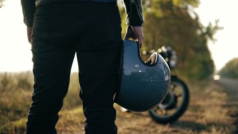 Back-view-of-and-unrecognizable-man-in-black-jeans-and-leather-jacket-holding-his-helmet-in-his-hand-while-standing-by-his-cool